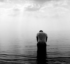 Black and white photo of a man with head bowed standing in ocean